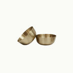 Two small metallic bronze bowls in a brushed brass or bronze finish, one standing upright and the other tilted on its side, shown against a plain off-white background.
