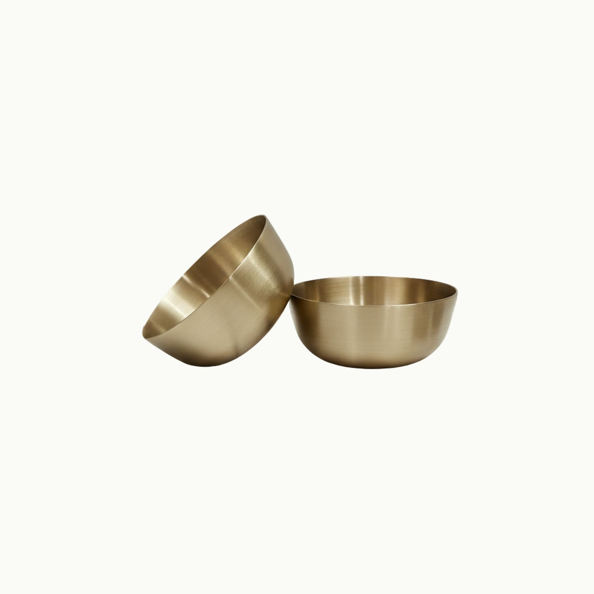 Two small metallic bronze bowls in a brushed brass or bronze finish, one standing upright and the other tilted on its side, shown against a plain off-white background.