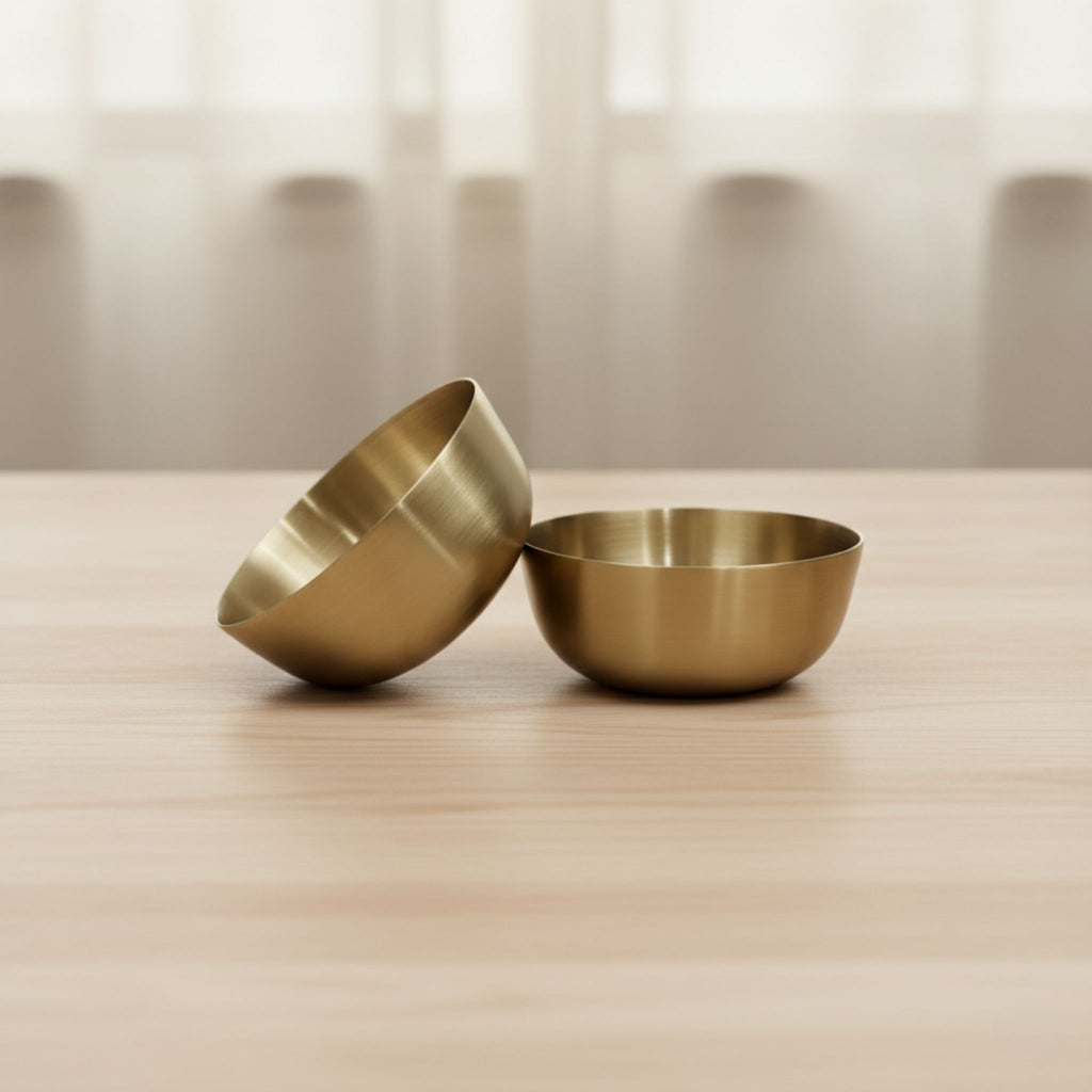 Two small metallic bronze bowls in a brushed brass or gold finish, one standing upright and the other tilted on a light wood surface with a softly blurred neutral background.
