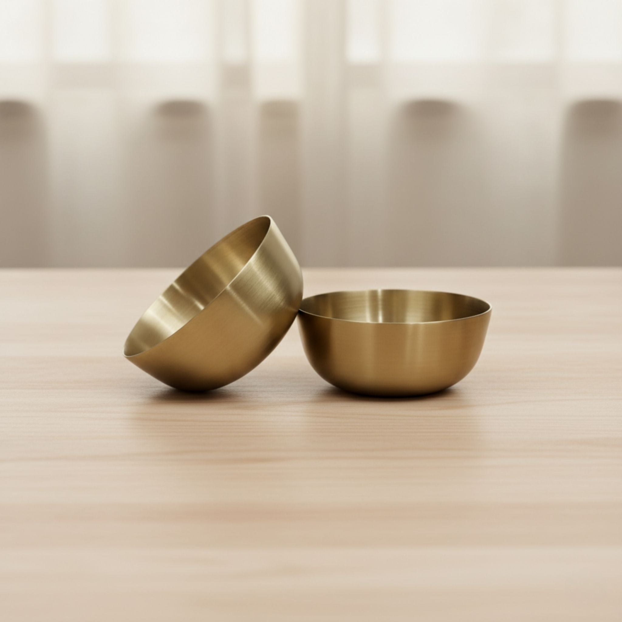 Two small metallic bronze bowls in a brushed brass or gold finish, one standing upright and the other tilted on a light wood surface with a softly blurred neutral background.