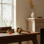 A brass bell-shaped incense burner with a curved handle lies on a wooden table in a softly lit room, alongside a reed diffuser and stacked books near a window.