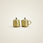 Two small metallic Brass teapot-style containers with lids and rounded handles, shown side by side on a plain warm off-white background.
