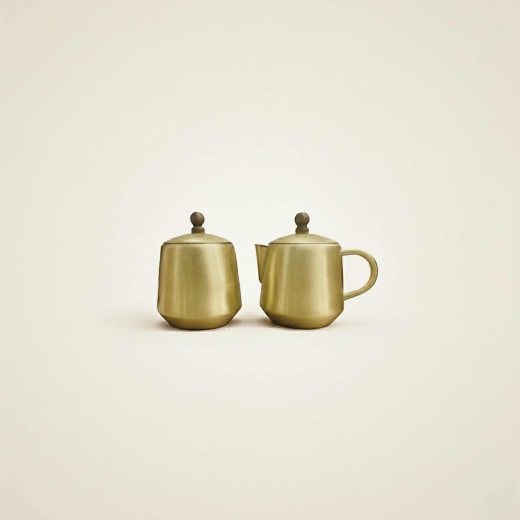 Two small metallic Brass teapot-style containers with lids and rounded handles, shown side by side on a plain warm off-white background.