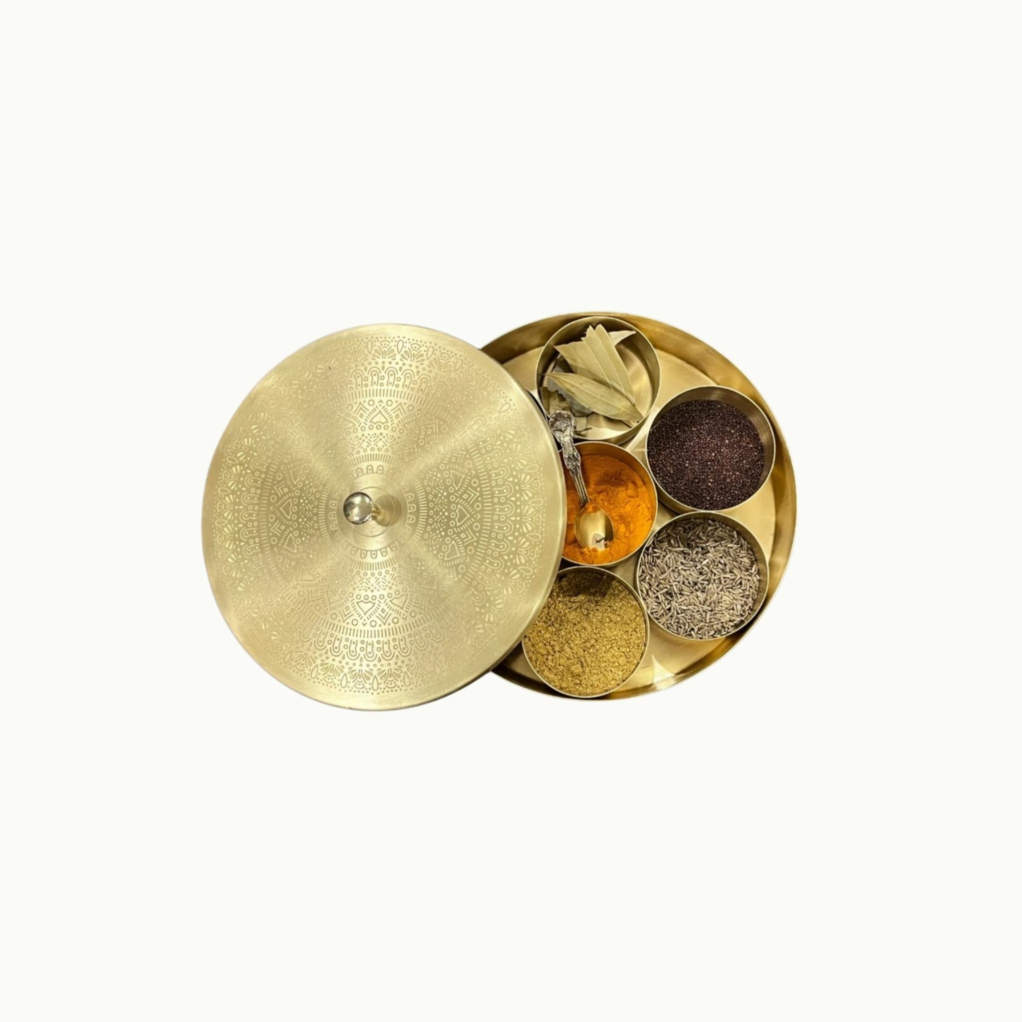 A brass Indian spice box with its lid off, showing six compartments filled with various whole spices and herbs, set against a plain off-white background.