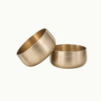 Two brushed bronze bowls, one tilted and one upright, on a clean off-white background.