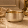 A brushed bronze metal bowl with concentric grooved details, set on a beige marble surface with draped satin fabric and a decorative tray in the background.