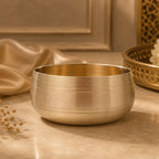 A brushed bronze metal bowl with concentric grooved details, set on a beige marble surface with draped satin fabric and a decorative tray in the background.