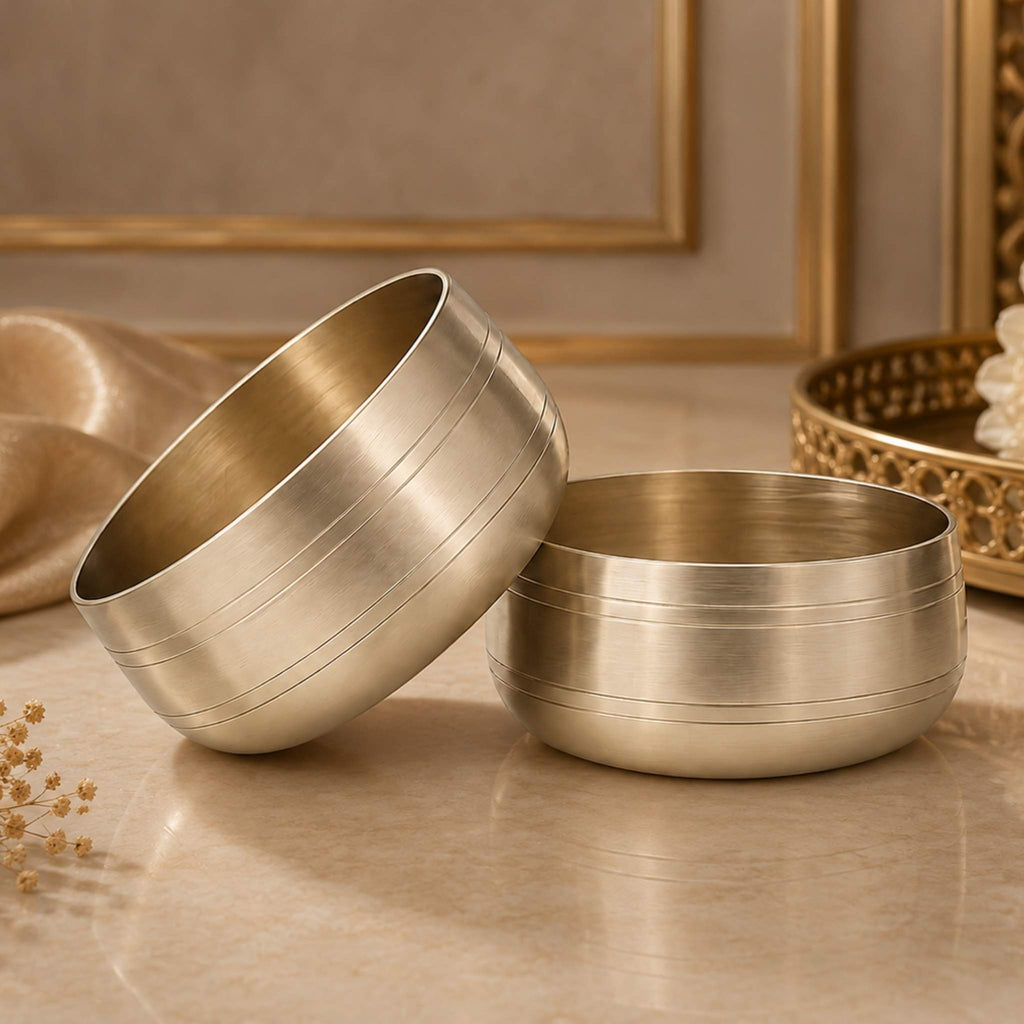 Two brushed metallic Bronze bowls with a warm gold finish, stacked slightly offset on a polished stone surface with a softly blurred decorative background.