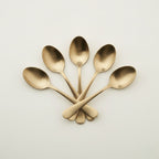 Five polished bronze metal spoons arranged in a fan shape with crossed handles on a clean, light off-white background.