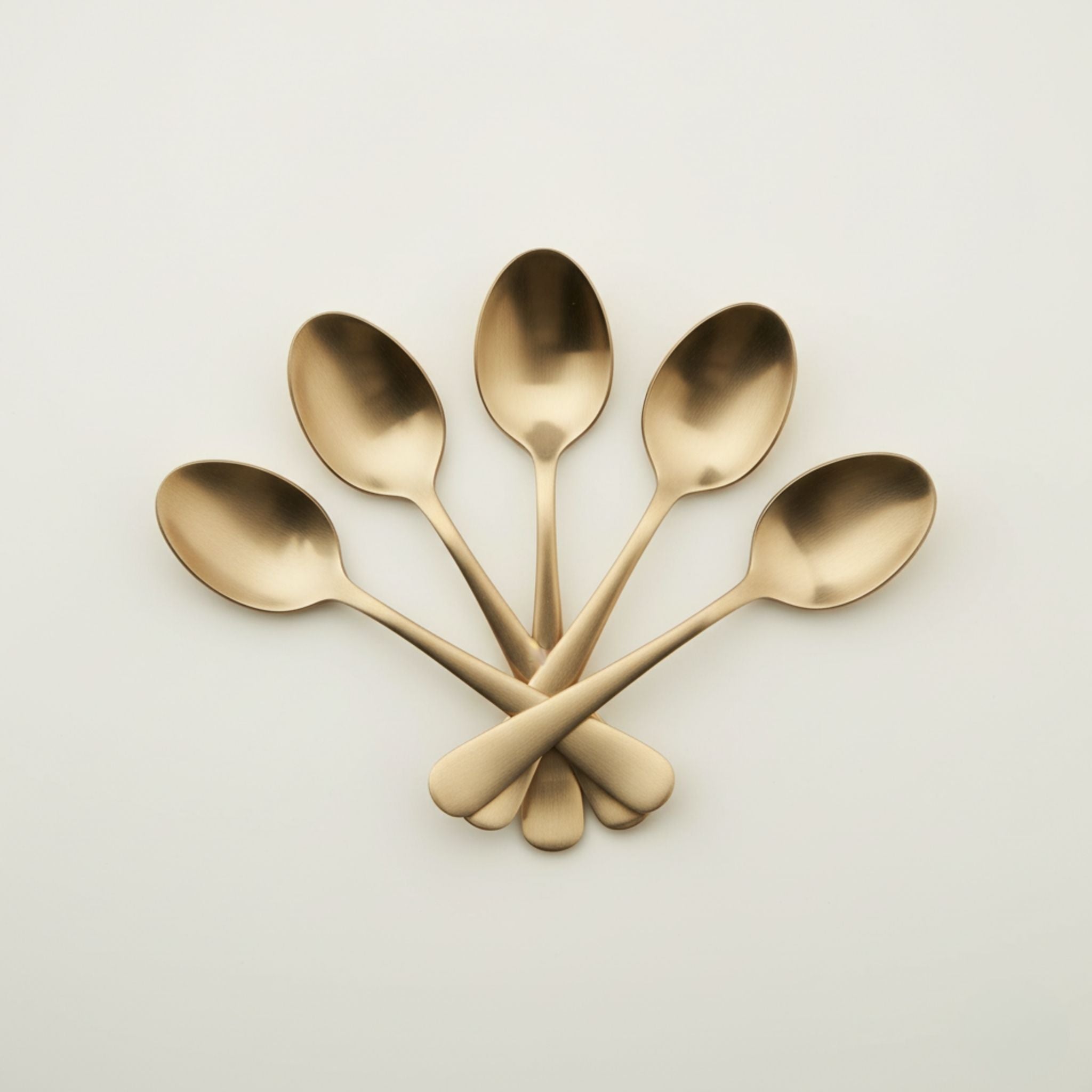 Five polished bronze metal spoons arranged in a fan shape with crossed handles on a clean, light off-white background.