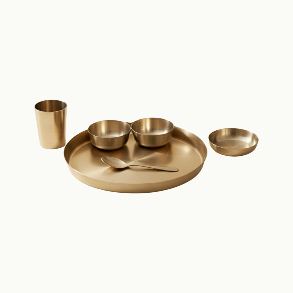 A minimalist bronze tableware set on a warm cream background, featuring a large oval tray with a spoon, two small bowls, a tall cup, and a shallow dish.