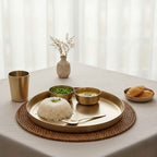 A styled tabletop bronze dining scene with a round gold-toned tray holding a mound of white rice, two small bowls (one with green peas, one with curry), a spoon, a small cup, and a plate with bread, arranged on a woven placemat on a beige tablecloth. A small vase with dried stems sits in the background near sheer curtains.