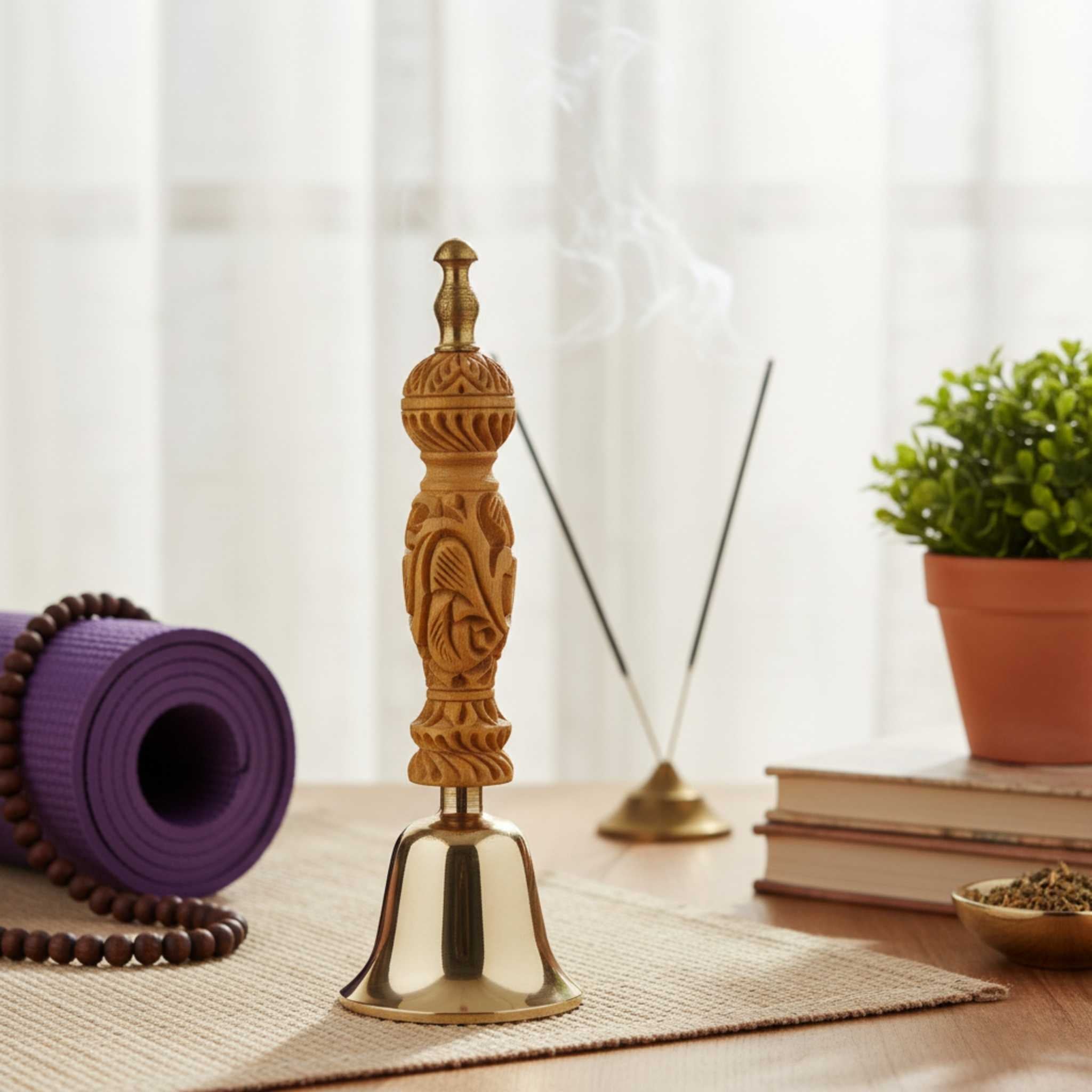 An ornate carved wooden incense burner with a polished brass bell base, styled on a woven mat on a wooden table with a purple yoga mat, incense sticks, books, and a potted plant in the background.