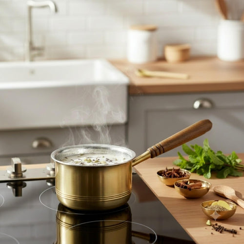 Brass saucepan with a wooden handle sits on a black induction cooktop in a bright kitchen, with steam rising from the soup inside. A wooden cutting board with fresh herbs and spices is visible on the right.