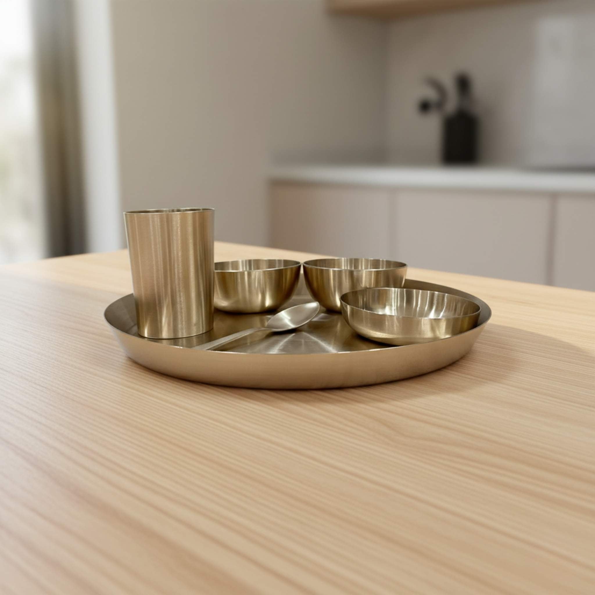 A bronze metal Indian thali set with a cup, three small bowls, and a spoon, arranged on a round tray on a light wood table.