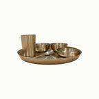 A bronze Indian food tray set with a large round platter, a tall cup, two small bowls, and a spoon, shown on a plain off-white background.