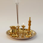 A polished brass ritual tray set with a incense holder holding two smoking incense sticks, a small oil lamp with a flame, a bowl, and a carved brass stand, all arranged on a round tray against a neutral light gray background.