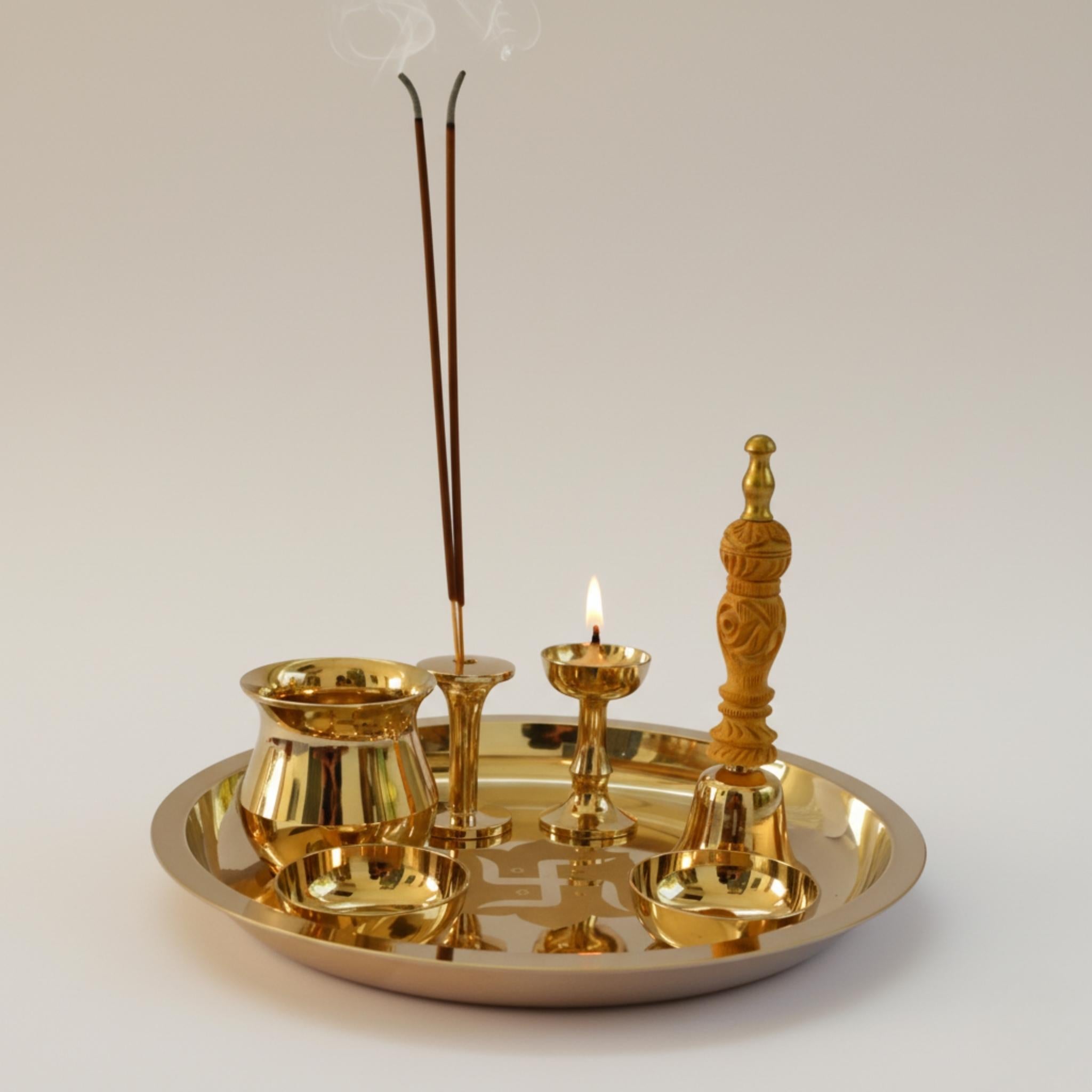 A polished brass ritual tray set with a incense holder holding two smoking incense sticks, a small oil lamp with a flame, a bowl, and a carved brass stand, all arranged on a round tray against a neutral light gray background.