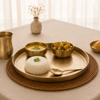 A bronze serving tray set on a woven placemat on a beige tablecloth, styled with a mound of white rice, a bowl of yellow curry, and a bowl of vegetable curry, alongside matching bronze cups and a spoon.